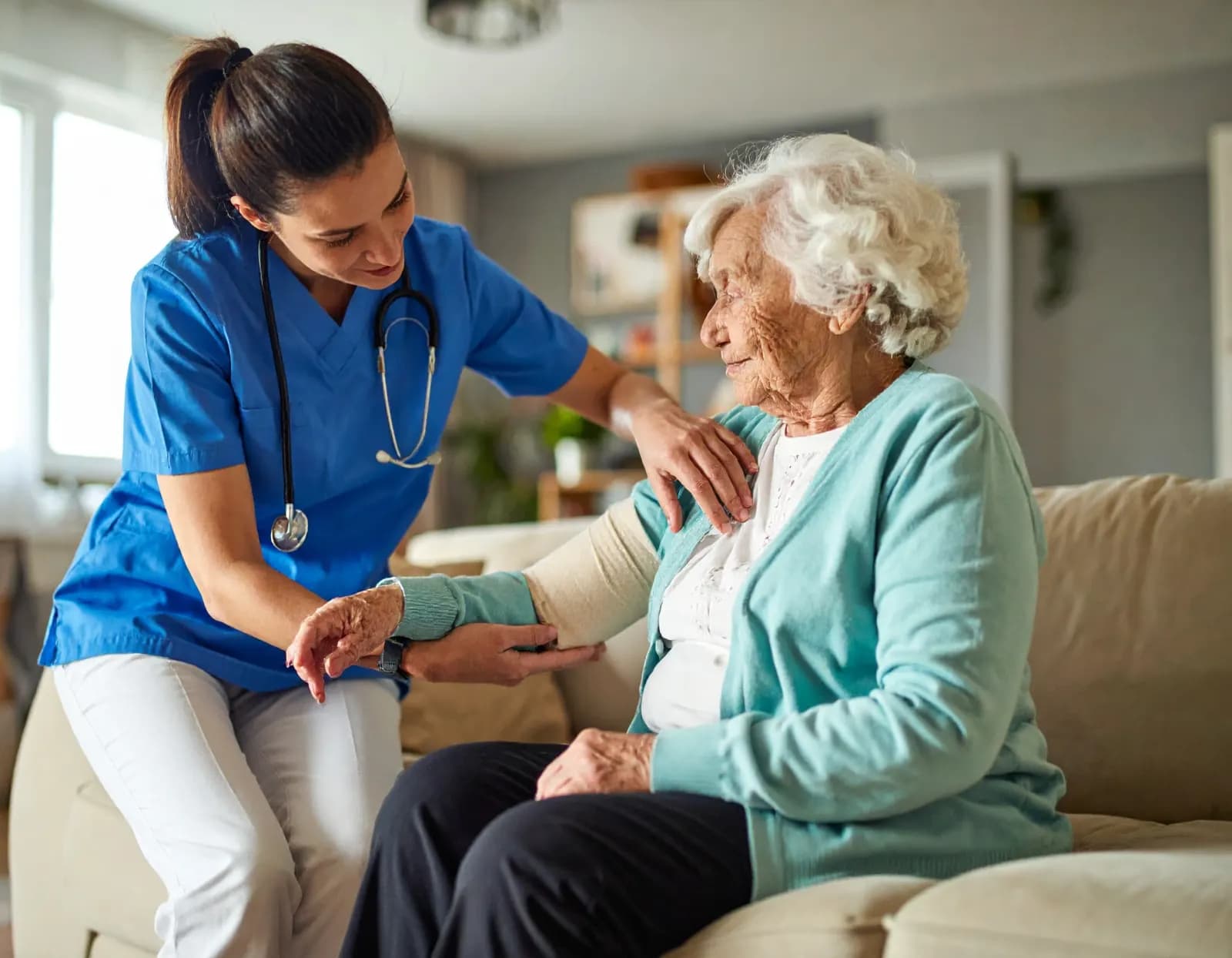 Asistentă medicală care o sprijină pe o femeie vârstnică acasă, pe canapea