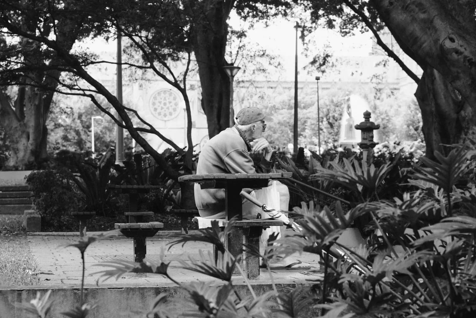 Vârstnic așezat la o masă de piatră într-un parc umbrit, fotografie alb-negru