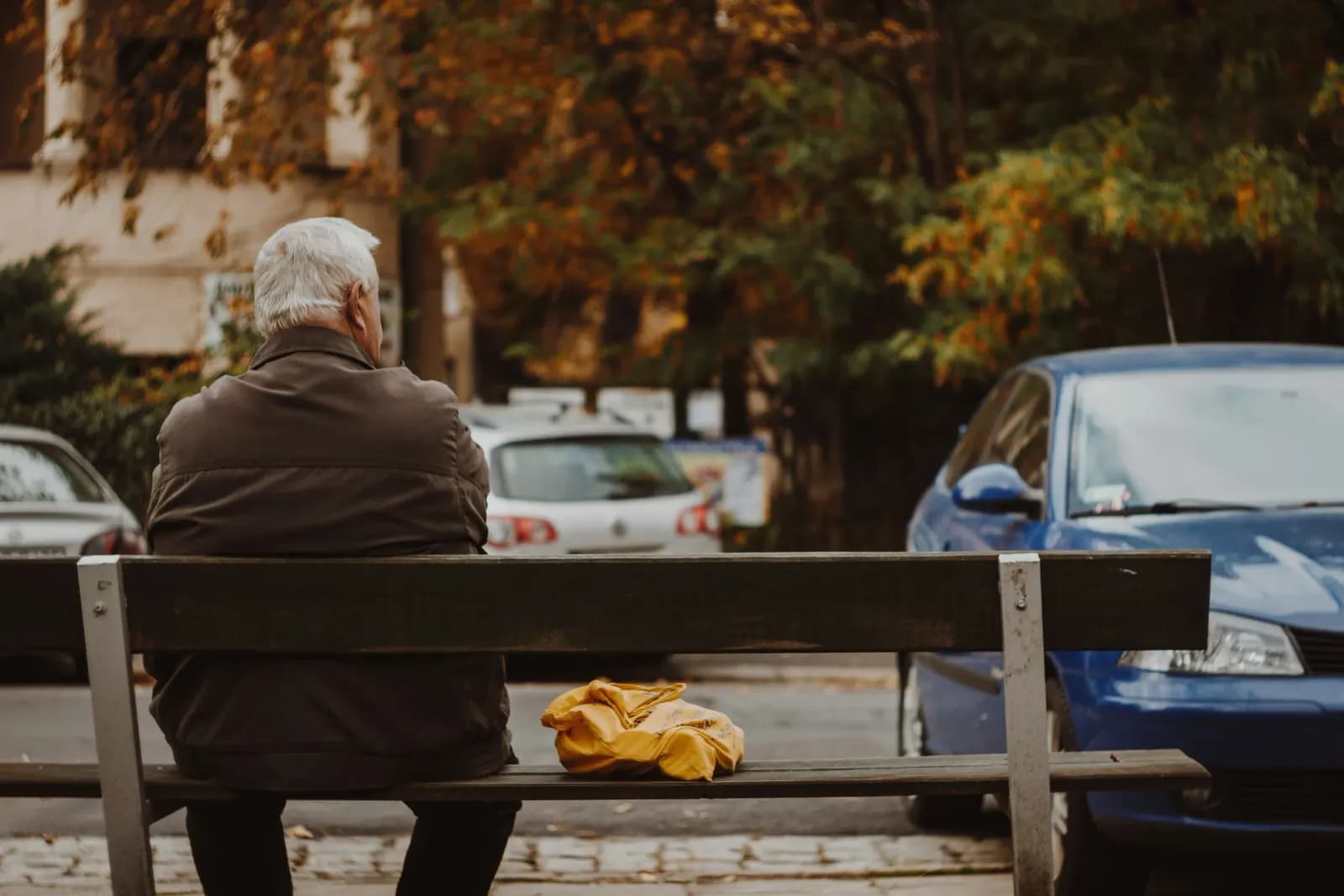 Vârstnic așezat singur pe o bancă, pe o stradă de toamnă