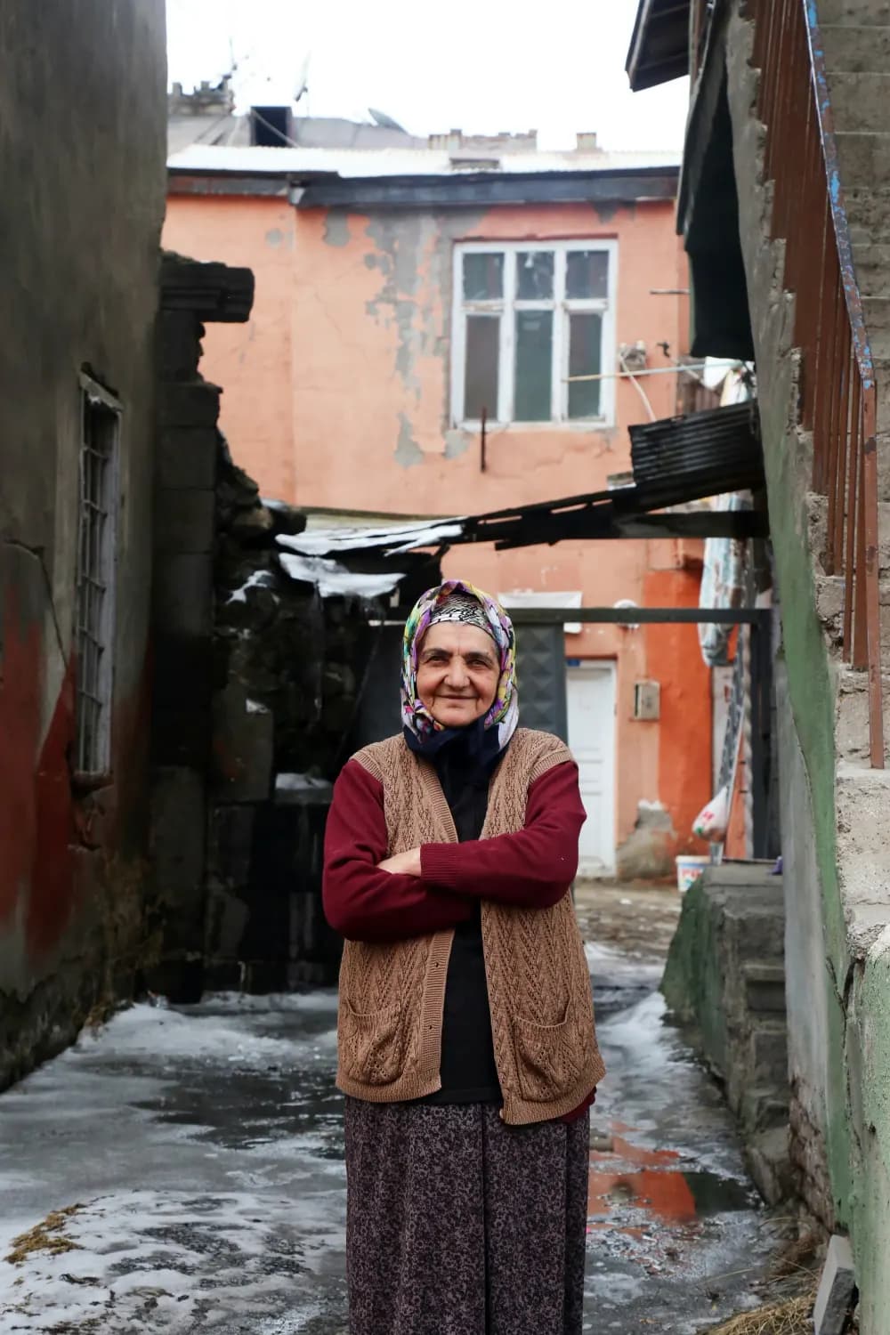 O doamnă în vârstă, cu basma, în fața casei ei
